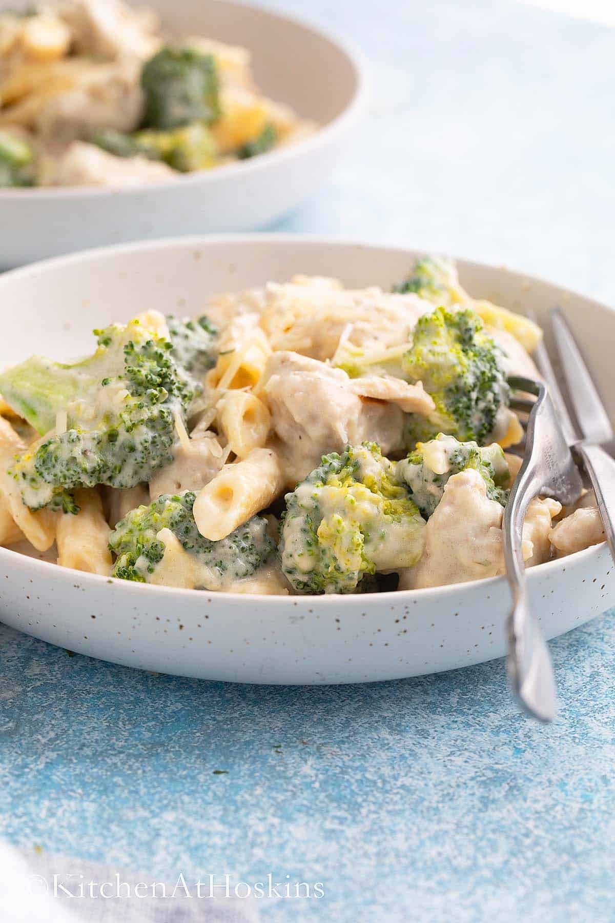 white plate with cooked pasta, chicken, broccoli and two forks.