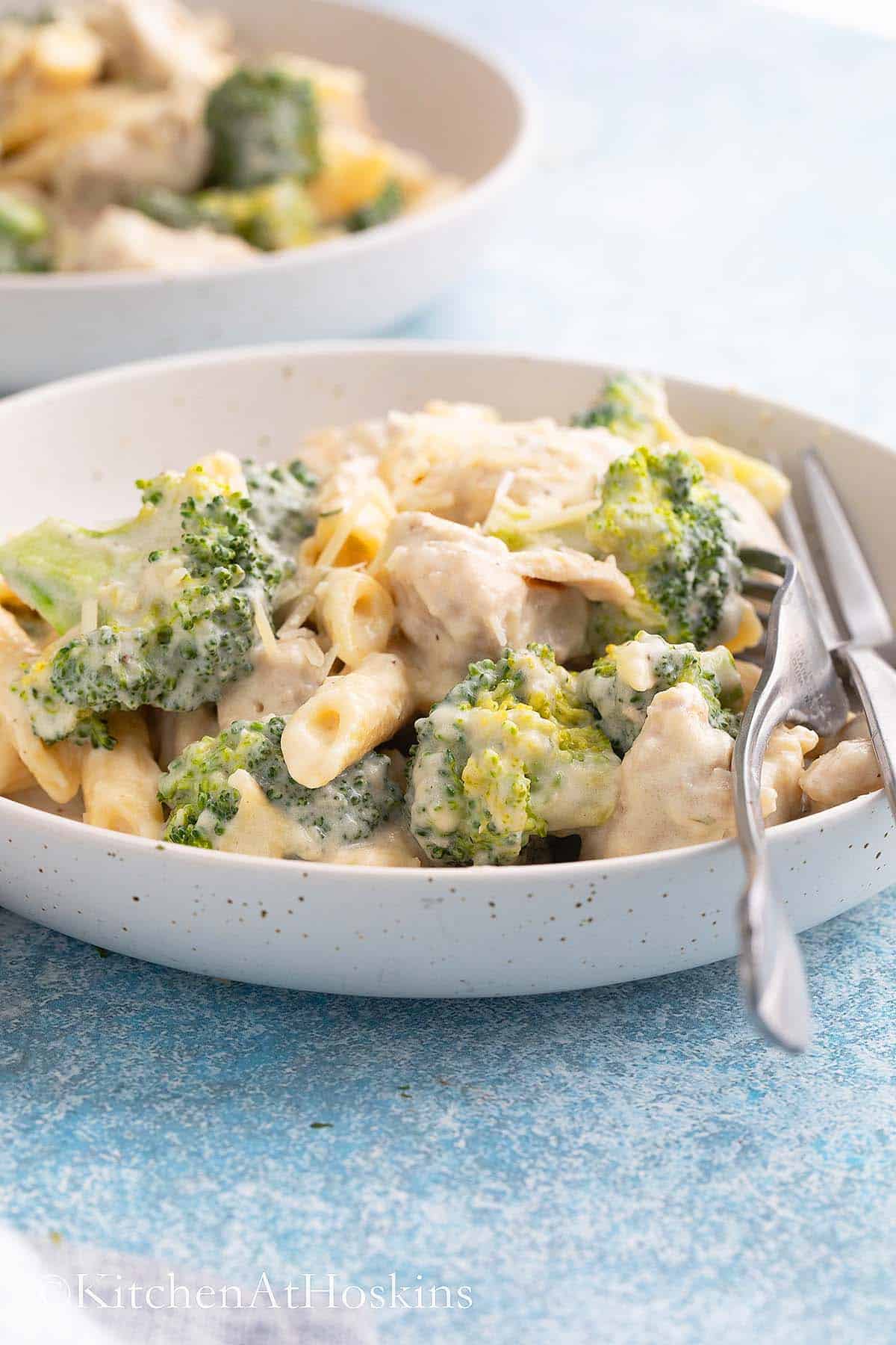 white plate with cooked pasta, chicken, broccoli and two forks.