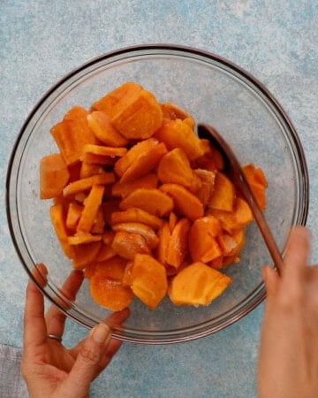 two hands mixing sliced persimmons in a large glass bowl.