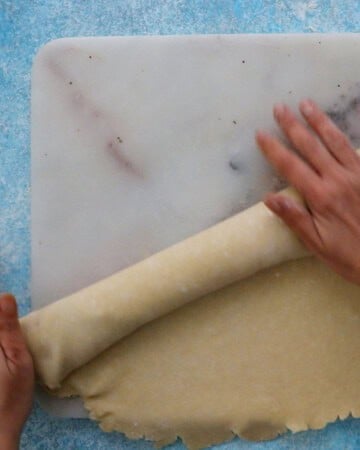 two hands rolling one pie crust on a marble board.