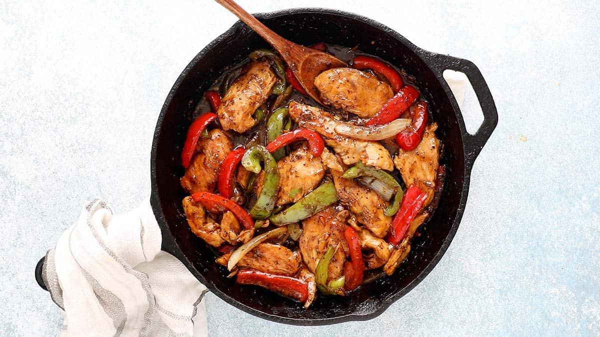 cooked black pepper chicken stir fry in a black skillet along with a wooden spoon.