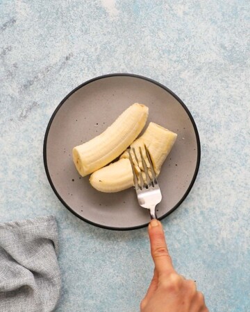 a hand mashing 2 bananas placed on a grey plate using a fork. 
