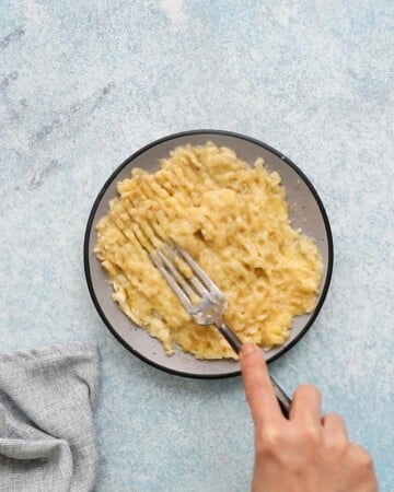 a hand mashing 2 bananas placed on a grey plate using a fork. 