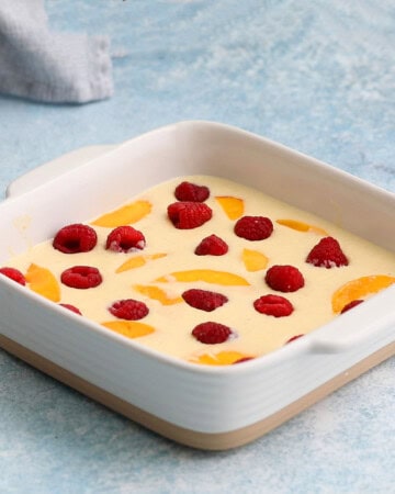 clafoutis batter topped with sliced mango and red raspberries in a square baking dish.