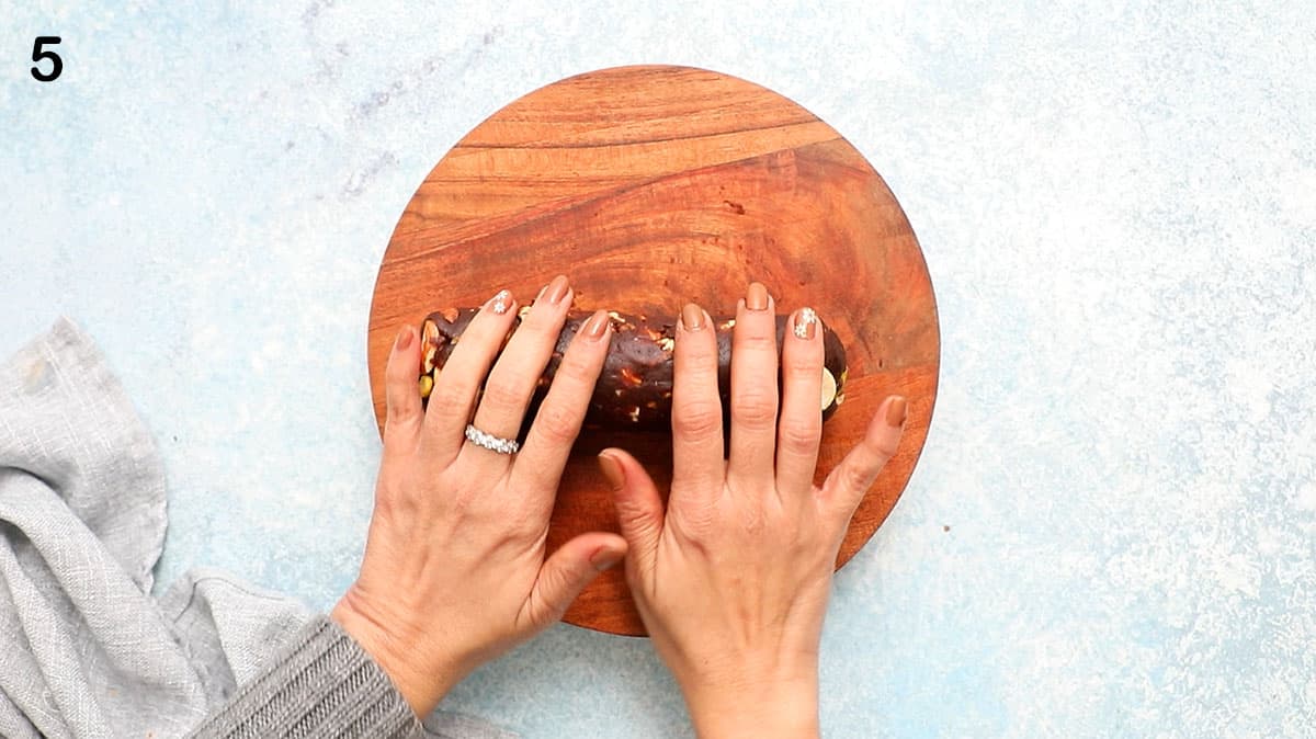 2 hands rolling a chocolate salami log on a wooden cutting board.