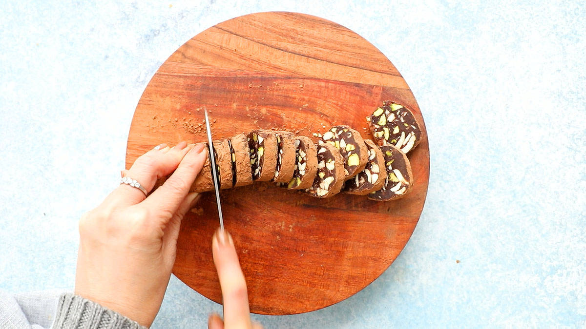 2 hands slicing a chocolate salami log into slices using a knife.