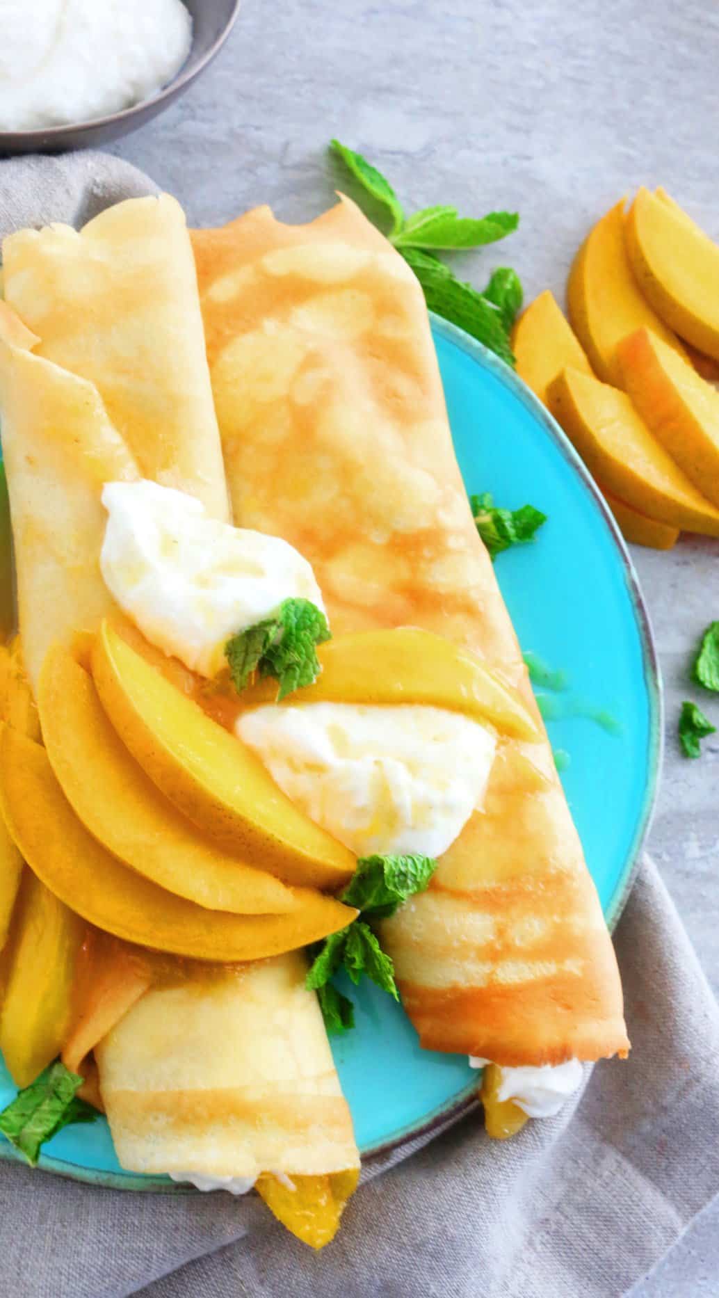 two rolled crepes topped with sliced mango on a blue plate.