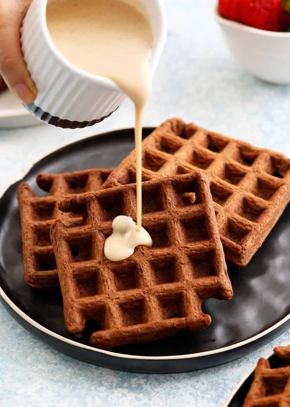 a hand pouring white sauce on top of three brown chocolate waffles placed on a black plate.