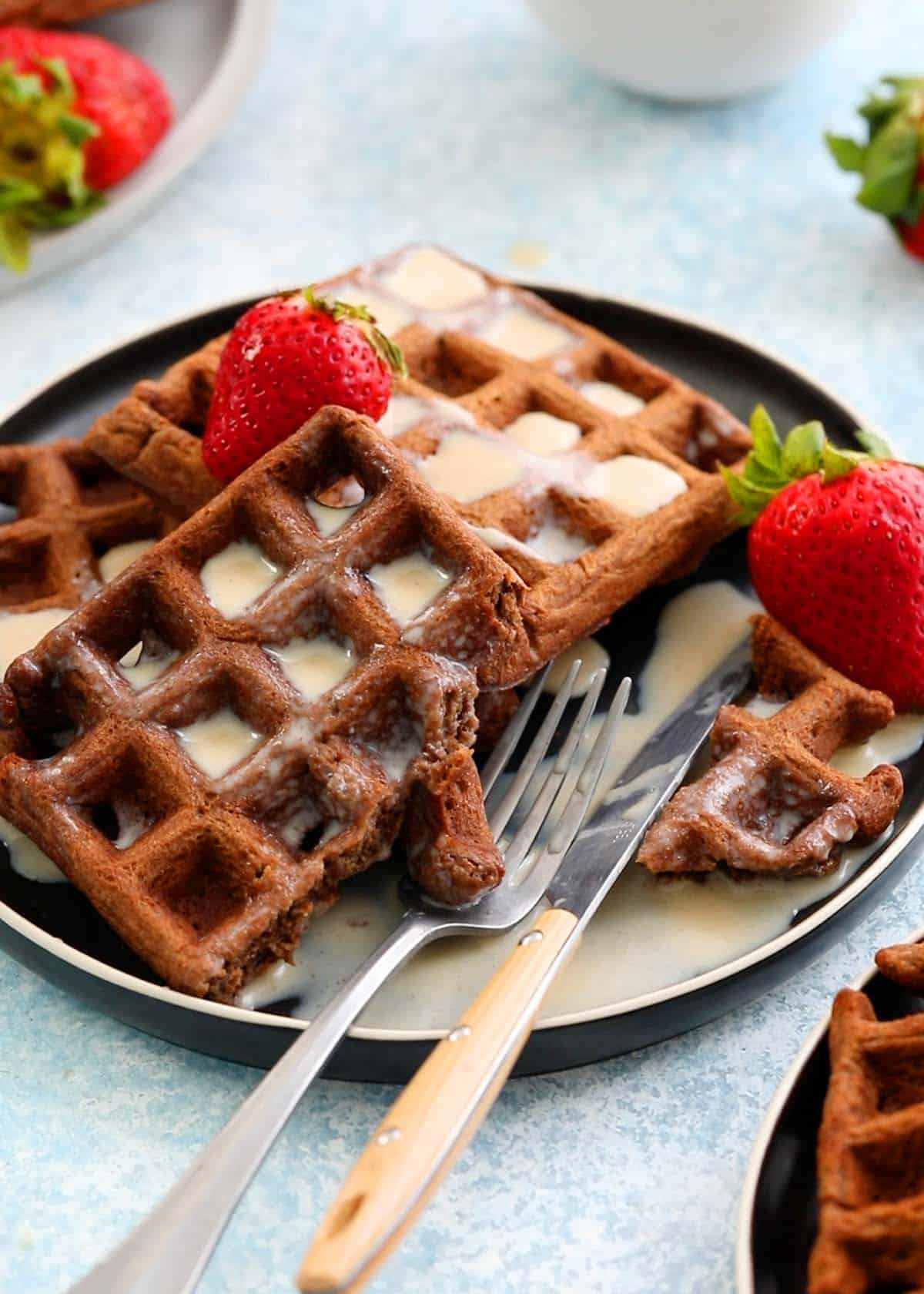 three brown chocolate waffles along with a fork and knife on a round black plate.