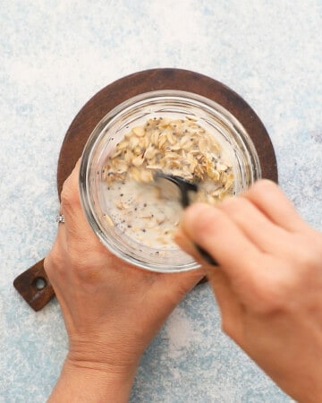 two hands stirring oats and milk in a glass jar.