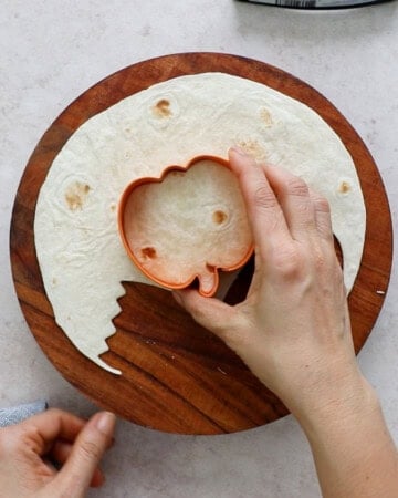 two hands cutting a white tortilla using a orange pumpkin cookie cutter.