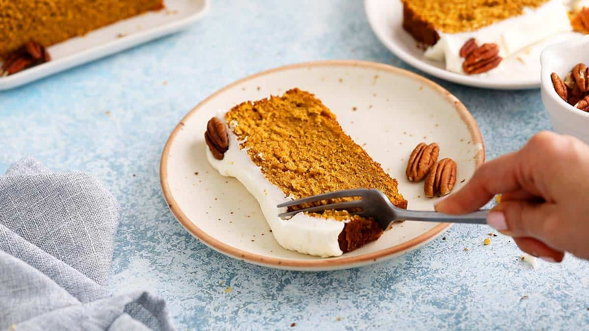 a hand using a fork to cut into a slice of pumpkin cake on a white plate.