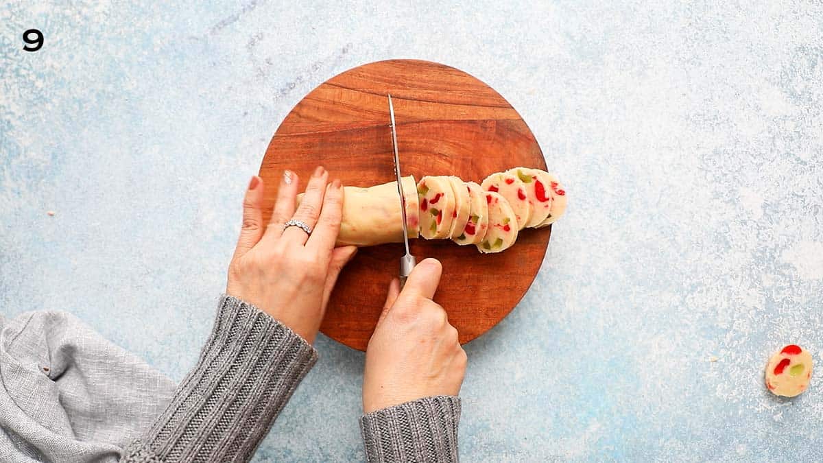 2 hands cutting a cookie dough log into slices using a knife.