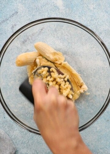 a hand mashing bananas in a glass bowl.