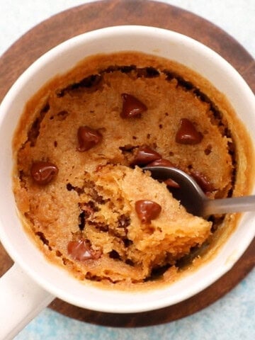 brown cake topped with chocolate chips in a white mug along with a spoon.