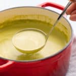 lifting a ladle with spinach soup from a red pot with the same.