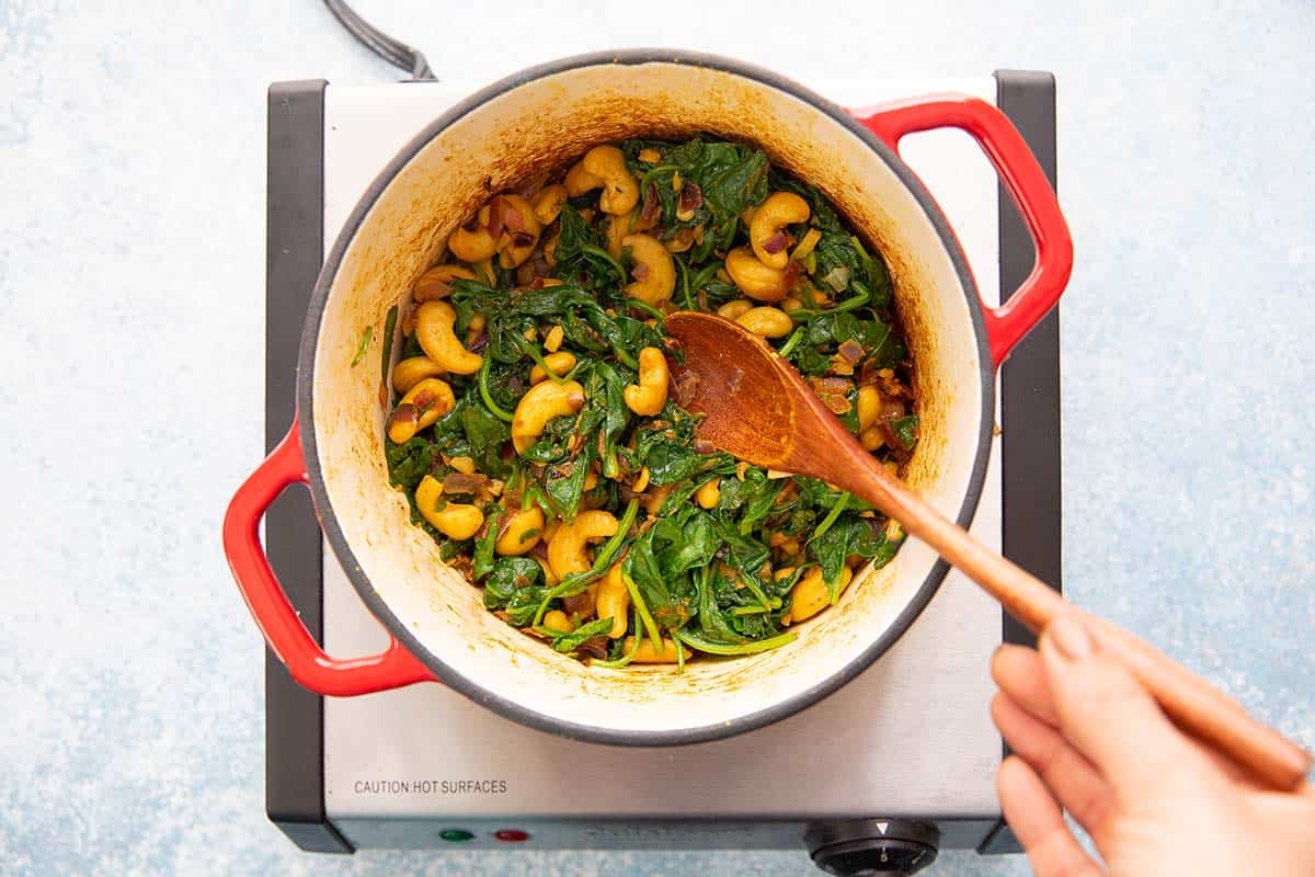 sauteing spinach, cashews, spices in a pot with a wooden spoon.
