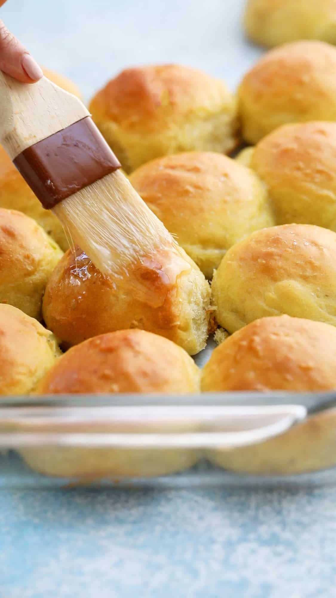 a hand brushing melted butter on baked dinner rolls.