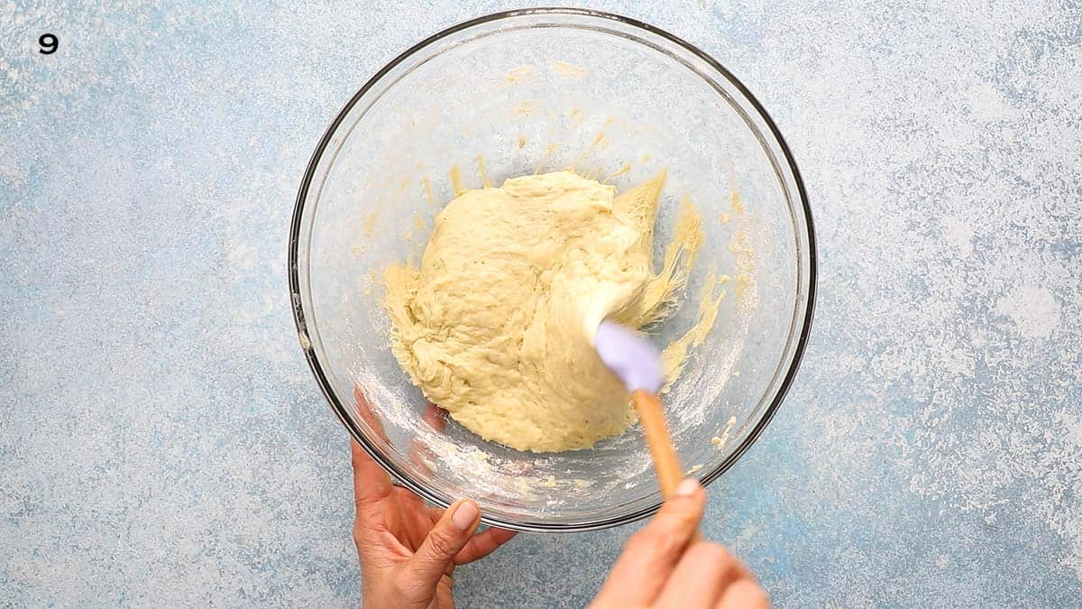 2 hands removing a risen yeast dough in a glass bowl with a violet spatula.