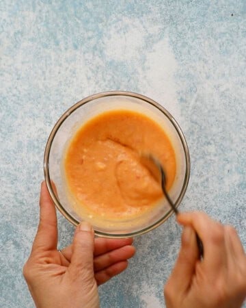 two hands mixing orange bang bang sauce in a small glass bowl.