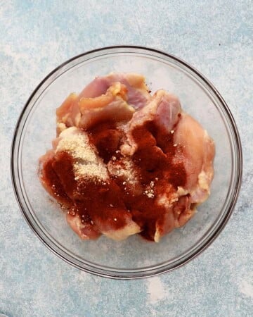 red seasonings on top of raw chicken in a glass bowl.