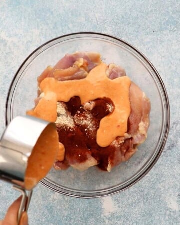 a hand pouring orange sauce over raw pink chicken thighs placed in a glass bowl.