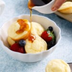 a hand pouring maple syrup over 4 pancake cups in a white bowl.