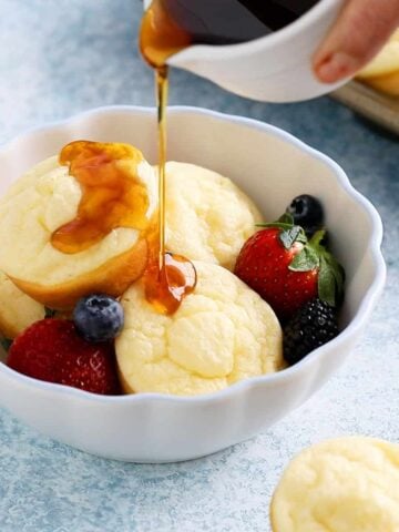 a hand pouring maple syrup over 4 pancake cups in a white bowl.