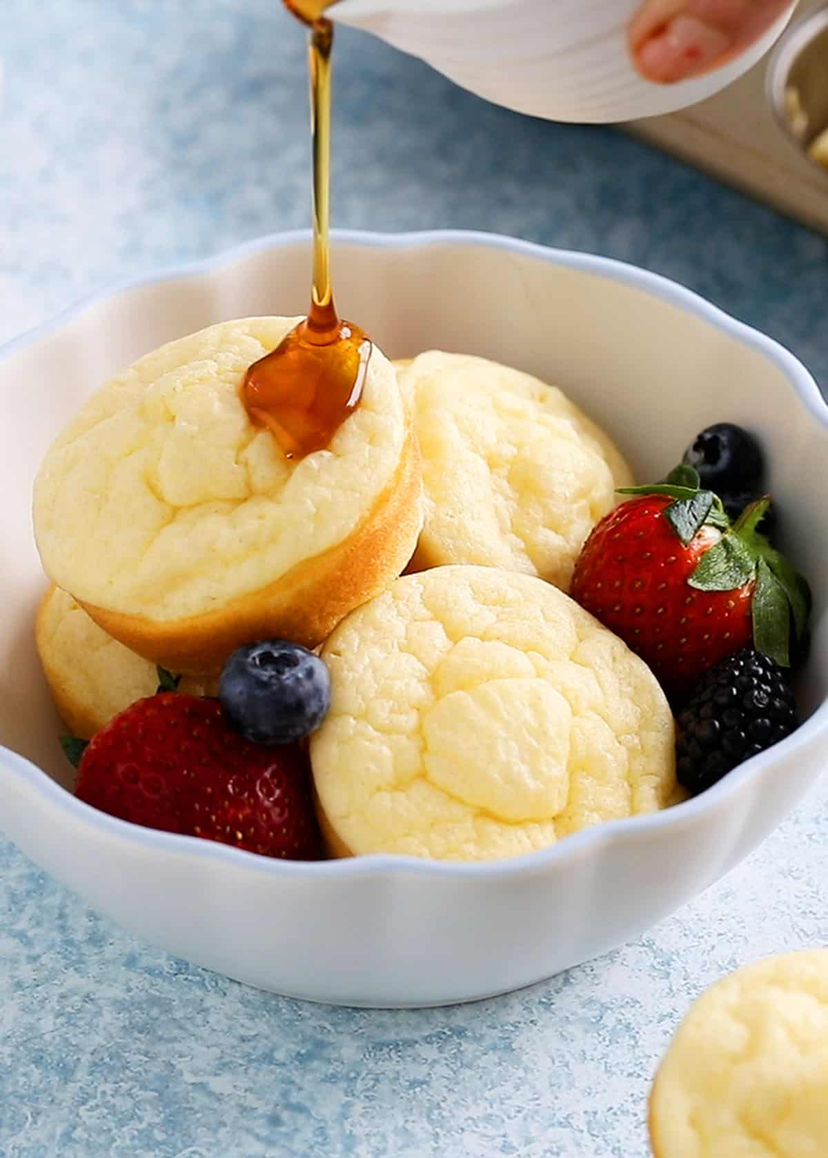 a hand pouring maple syrup over baked pancake muffins in a white bowl.