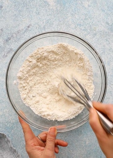 two hands mixing white flour in a glass bowl with a metal whisk.