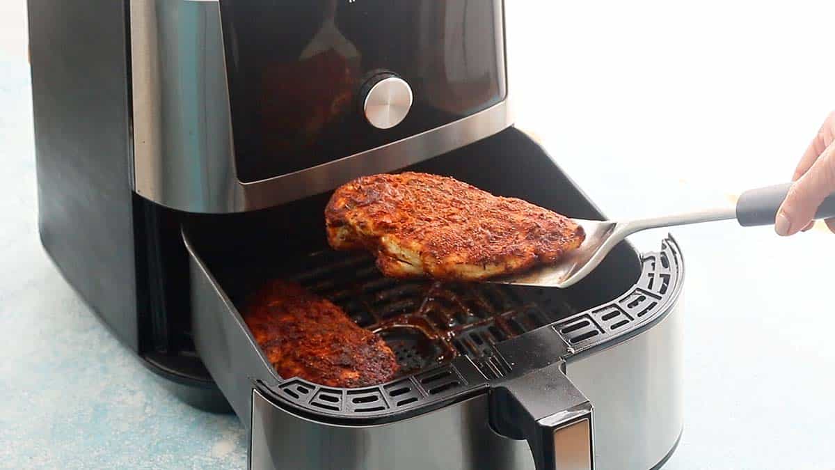 a hand lifting one cooked chicken breast from an air fryer.