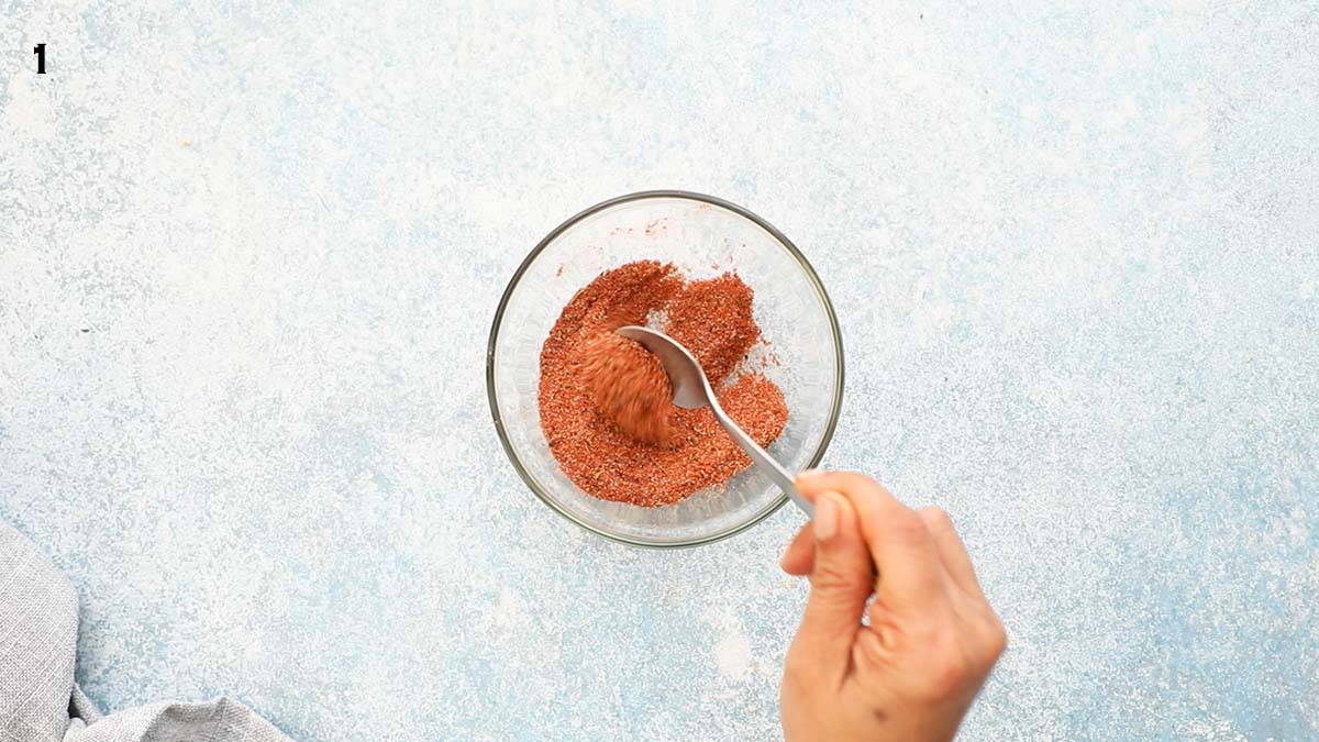 a hand mixing spices in a small glass bowl using a metal spoon.