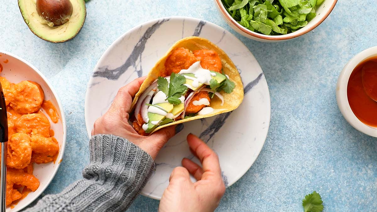 2 hands grabbing a shrimp taco from a white round plate.