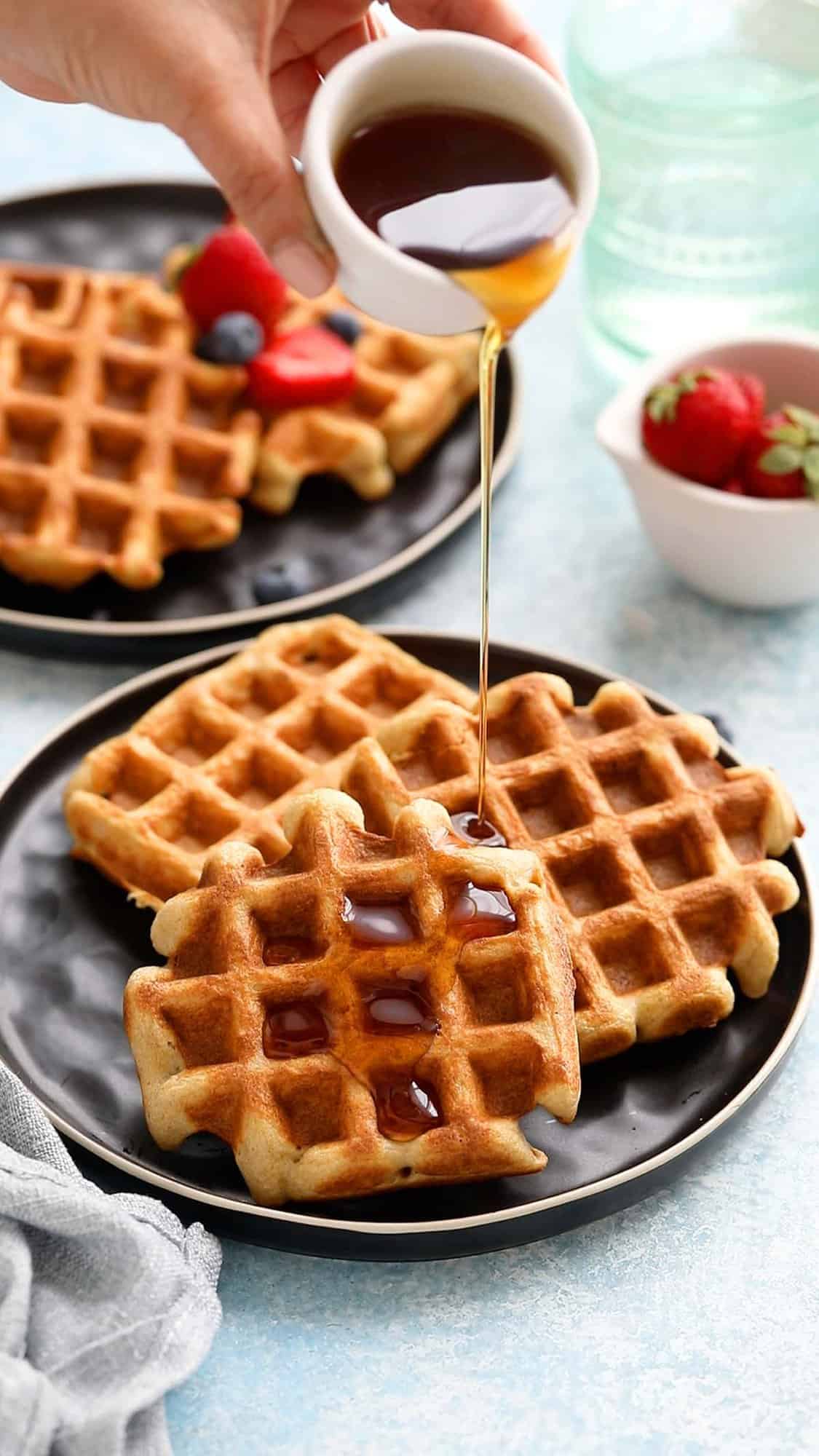 a hand pouring maple syrup on 3 three waffles placed on a black plate.