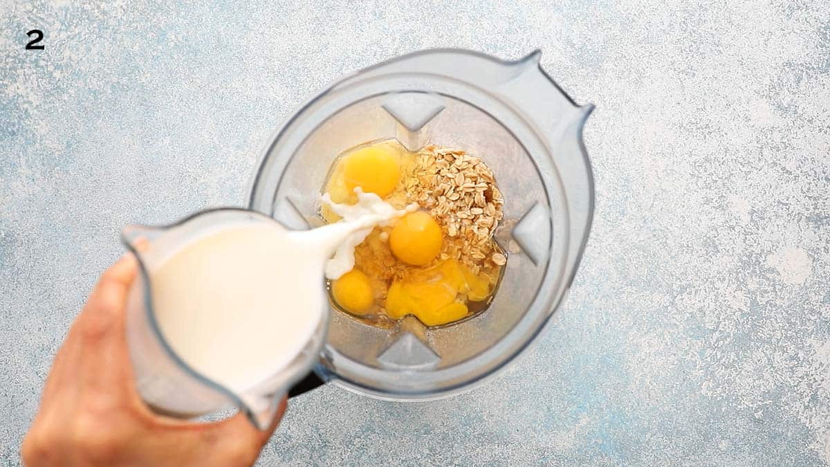 a hand pouring milk into a blender with dry ingredients.