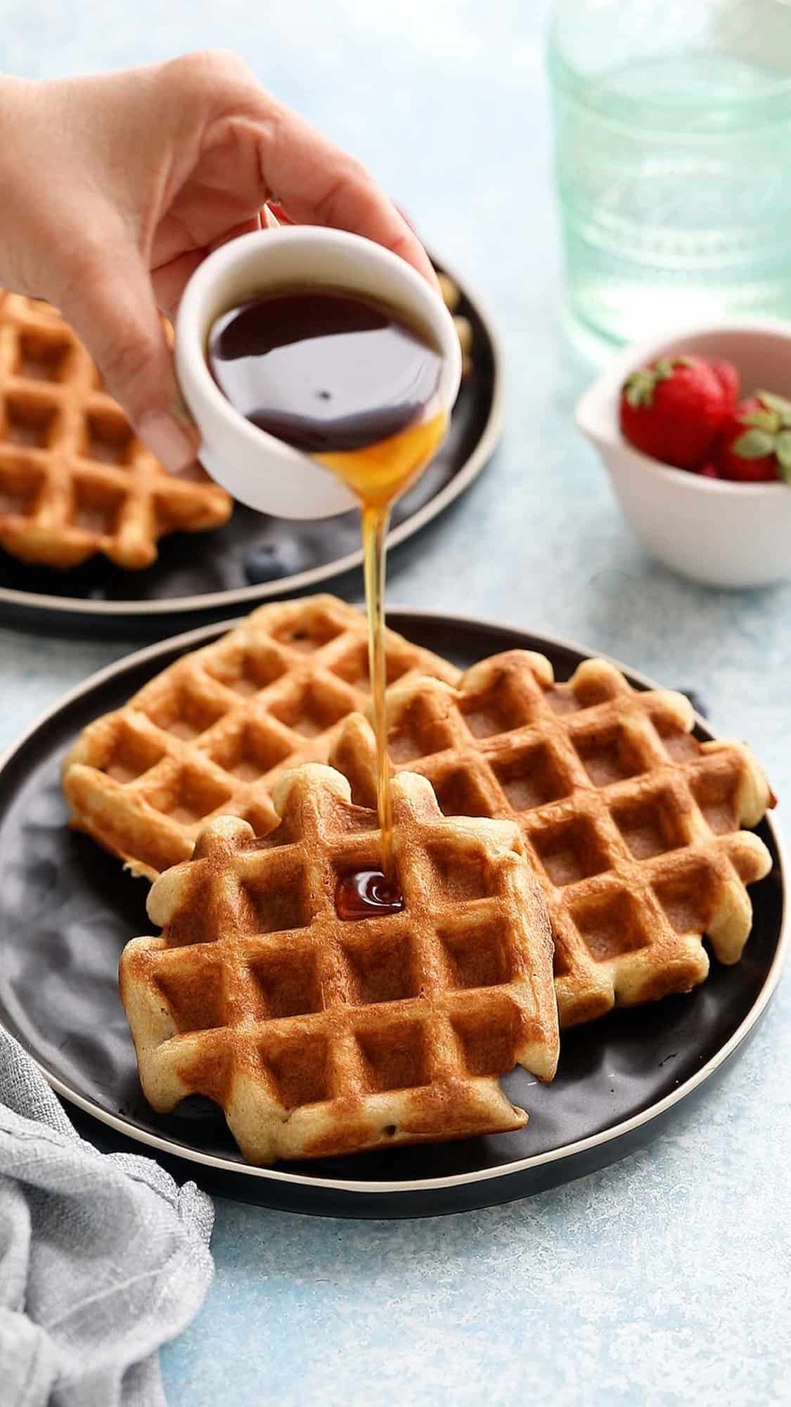 a hand pouring maple syrup on 3 three waffles placed on a black plate.
