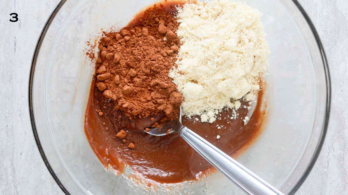 a stainless steel spoon in a glass bowl with melted chocolate, almond flour and cocoa powder.