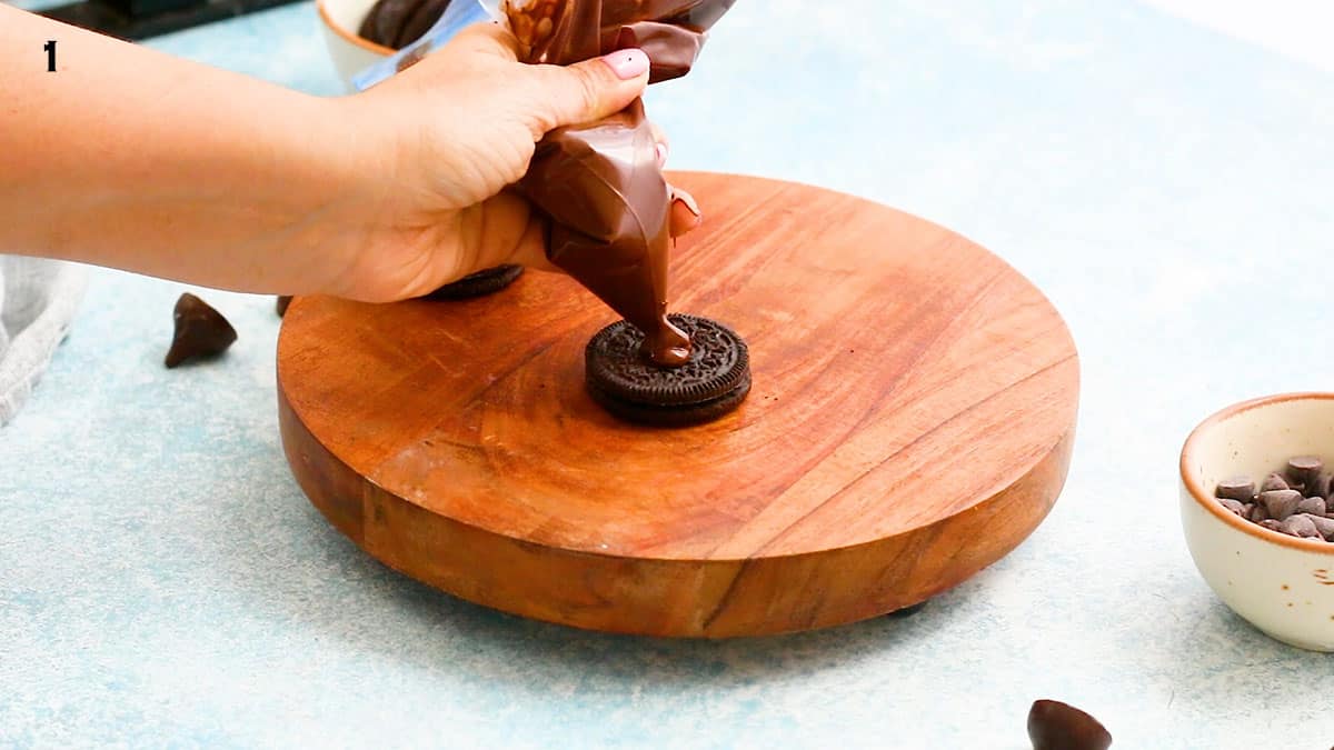 a hand squeezing melted chocolate on top of an Oreo cookie, using a pastry bag.