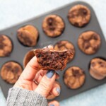 a hand holding half a chocolate muffin above a muffin pan.