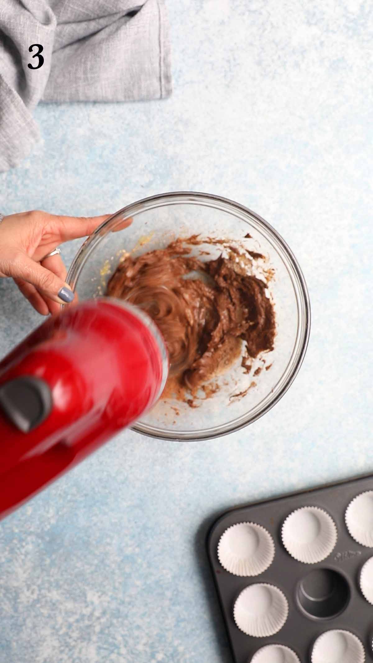 mixing chocolate muffin batter in a glass bowl with a red electric beater.