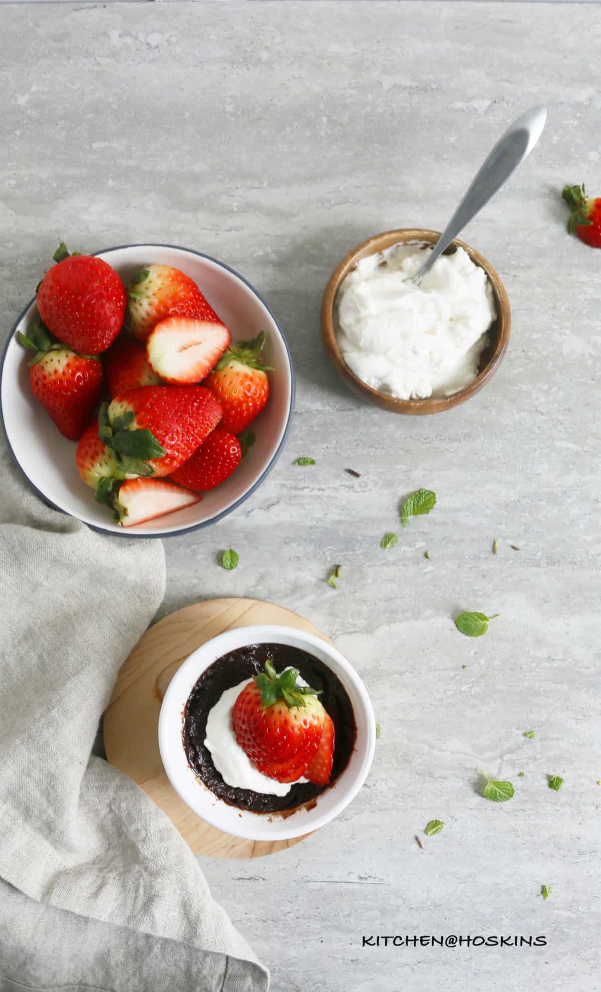 a white bowl with fresh strawberries, a wooden bowl with whipped cream along with a spoon and a white ramekin with fudgy nutella brownie