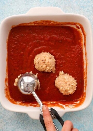 a hand scooping chicken meatballs in a white dish with red sauce.