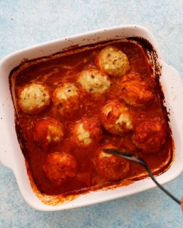 a hand mixing red sauce and chicken meatballs in a white baking dish.