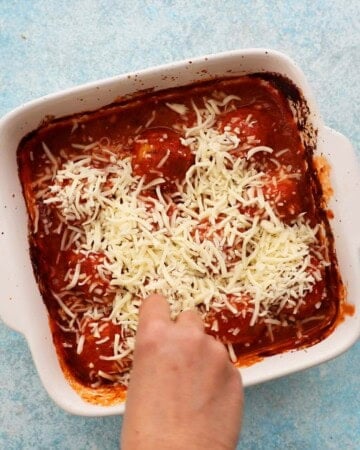 a hand adding white cheese into a white baking dish with red sauce.