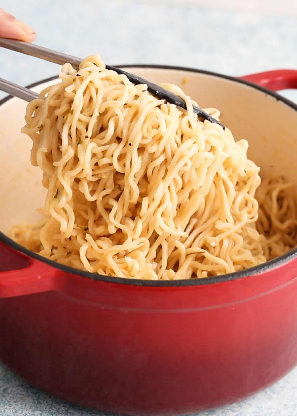 a hand removing cooked yellow ramen noodles from a red pot using black tongs.