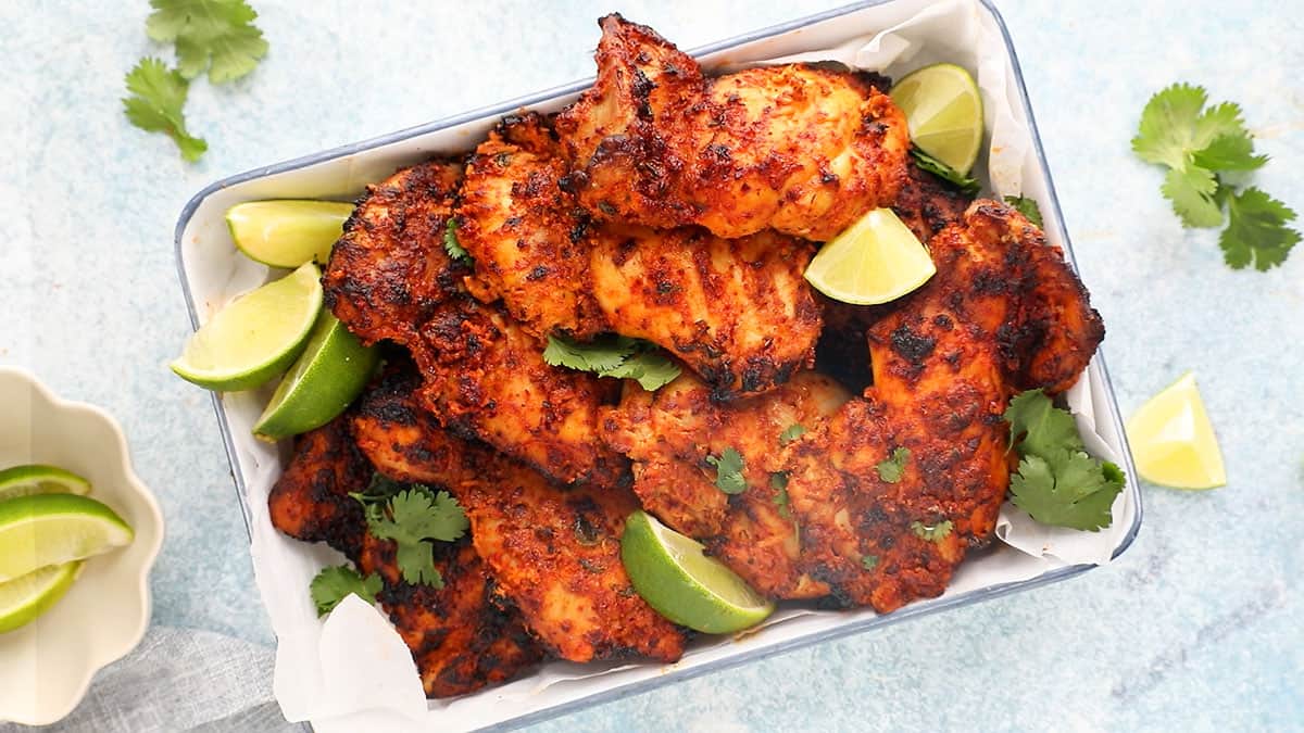 cooked tandoori chicken thighs placed in a rectangular white tray along with lime wedges.