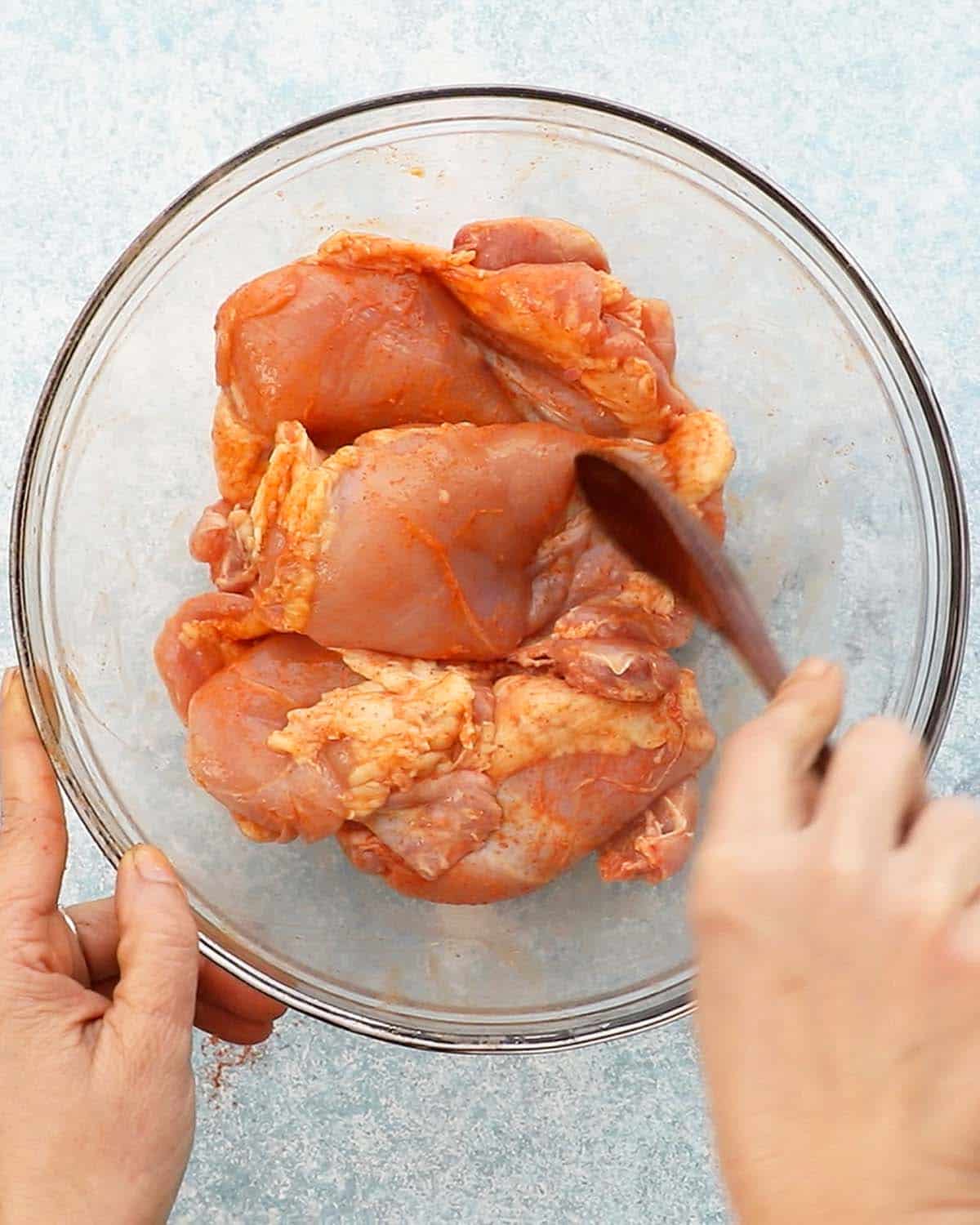 two hands tossing raw chicken thighs with seasoning in a glass bowl. 