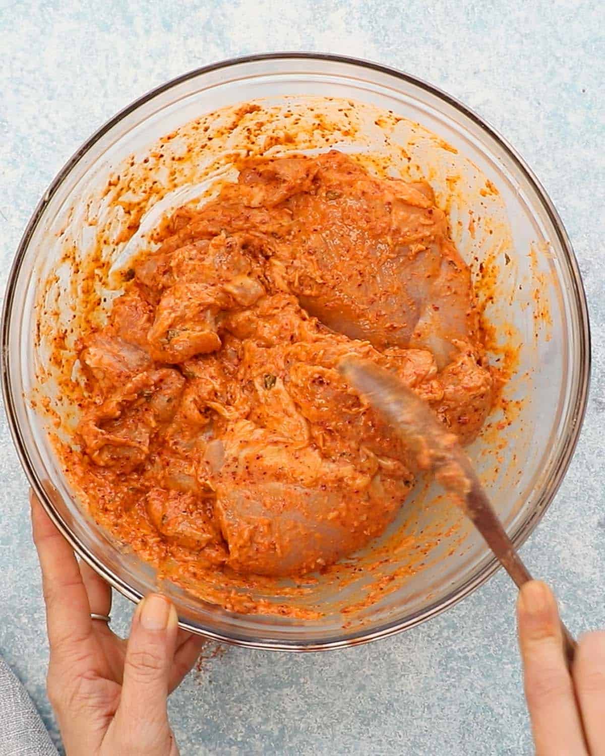 two hands mixing chicken thighs with orange marinade in a glass bowl.