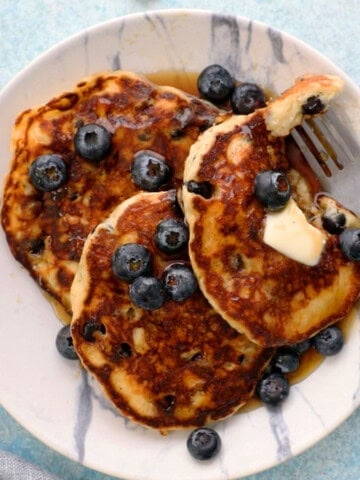 3 blueberry pancakes in a round white plate along with a fork.