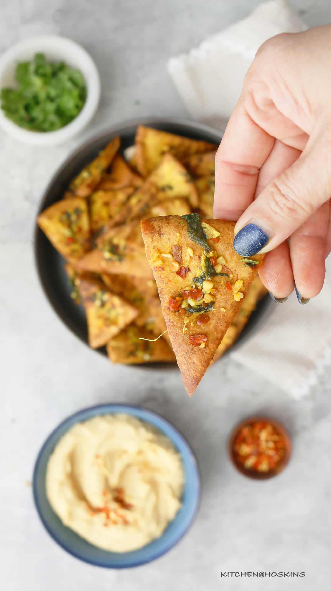 a hand holing a homemade baked pita chip topped with seasoning and herbs. 